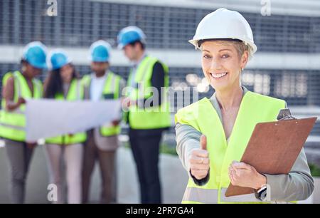 Wir haben dieses Projekt in kürzester Zeit abgeschlossen. Gekürztes Porträt einer attraktiven Bauarbeiterin, die Daumen nach oben zeigt, während sie mit ihren Kollegen im Hintergrund auf einer Baustelle steht. Stockfoto