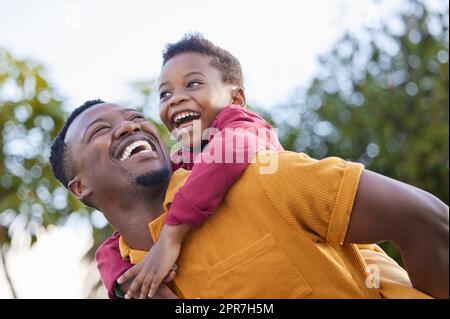 Ein entzückender kleiner Junge, der mit seinem Vater in einem Garten eine Huckepack-Fahrt genießt. Stockfoto