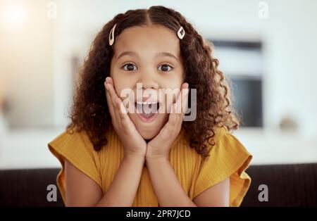 Das niedliche, gemischte Kind berührt zu Hause mit seinen Händen das Gesicht. Ein kleines süßes spanisches Mädchen, das allein auf einem Sofa im Wohnzimmer sitzt und schockiert aussieht. Aufgeregtes Kind mit lockigem Haar Stockfoto