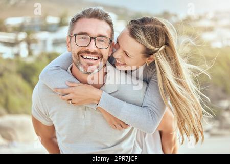 Glückliches und liebevolles, reifes, weißes Paar, das an einem sonnigen Tag ein romantisches Date am Strand zusammen genießt. Fröhlicher, liebevoller Ehemann, der seine Frau im Huckepack trägt, während er sich im Urlaub im Freien anfreundet Stockfoto