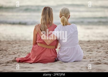 Rückansicht einer kaukasischen Mutter und Tochter, die am Strand zusammen auf dem Sand sitzen und die Aussicht betrachten Stockfoto