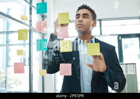 Ein seriöser kreativer Geschäftsmann aus gemischten Rassen, der allein auf der Arbeit Ideen für die Planung an einem Glasfenster schreibt. Ein fokussierter hispanischer Geschäftsmann, der in einem Büro Brainstorming durchführt Stockfoto