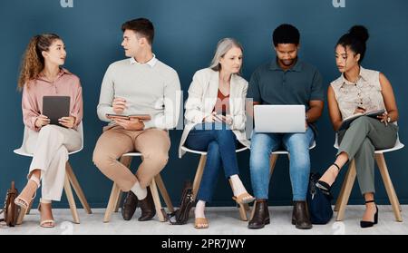 Eine Gruppe seriöser Geschäftsleute, die auf Stühlen in einer Reihe in einem Büro sitzen. Fünf Geschäftsleute sitzen und unterhalten sich mit Technologie in einem Wartezimmer in einem Büro Stockfoto