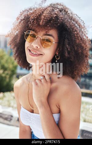 Porträt einer jungen, trendigen, wunderschönen Frau mit gemischter Rasse mit einem Afro, das lächelt und alleine draußen posiert. Eine hispanische Frau, die eine Sonnenbrille trägt und glücklich ist. Modische afroamerikanische Frau in der Stadt Stockfoto