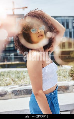 Porträt einer jungen, trendigen, wunderschönen Frau mit gemischter Rasse mit einem Afro, das lächelt und alleine draußen posiert. Eine hispanische Frau, die eine Sonnenbrille trägt und glücklich ist. Modische afroamerikanische Frau in der Stadt Stockfoto