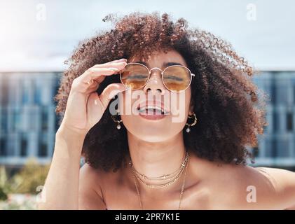 Junge Mischrasse mit lockigem, natürlichem Afrohaar und trendiger, stilvoller Sonnenbrille. Eine Frau sieht nur sorglos, cool und selbstbewusst aus. Fröhliche, modische hispanische Frau in der Stadt Stockfoto