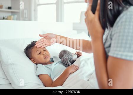 Ein kranker kleiner Junge im Bett, während seine Mutter telefoniert und einen Arzt um Hilfe bittet, um das Fieber zu senken. Junge alleinerziehende Eltern fühlen, dass Söhne auf der Stirn die Temperatur messen. Gemischtes Kind, das sich krank fühlt und im Bett liegt, während seine Mutter Fieber untersucht Stockfoto