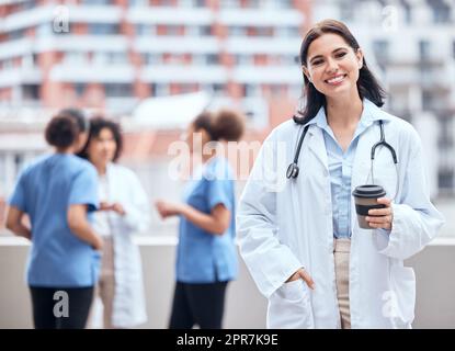 Porträt einer lächelnden jungen Ärztin, die draußen eine Tasse Kaffee mit ihren Kollegen im Hintergrund genießt. Glücklicher Arzt macht eine Pause von der Arbeit im Krankenhaus, um Tee zu trinken. Stockfoto