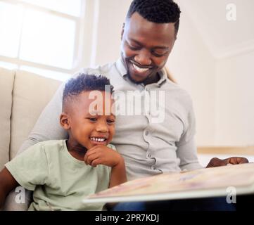 afroamerikanischer Vater, der sich mit seinem zu Hause anfreundet. Schwarzer Mann, der seinem Sohn hilft, ein Buch zu lesen und das Lernen zu üben, während er zu Hause auf dem Sofa sitzt. Süßer kleiner schwarzer Junge, der seinem Vater im Wohnzimmer vorliest. Vater und Sohn lächeln, während sie etwas lehrreich lernten Stockfoto