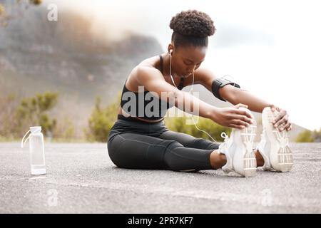 Eine afroamerikanische Sportlerin mit einem Afro, die Musik auf ihren Kopfhörern hört, während sie draußen in der Natur trainiert. Eine engagierte schwarze Frau lächelt, während sie sich aufwärmt, bevor sie draußen trainiert Stockfoto