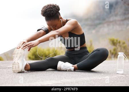 Eine afroamerikanische Sportlerin mit einem Afro, die Musik auf ihren Kopfhörern hört, während sie draußen in der Natur trainiert. Eine engagierte schwarze Frau lächelt, während sie sich aufwärmt, bevor sie draußen trainiert Stockfoto