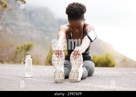 Eine afroamerikanische Sportlerin mit einem Afro, die Musik auf ihren Kopfhörern hört, während sie draußen in der Natur trainiert. Eine engagierte schwarze Frau lächelt, während sie sich aufwärmt, bevor sie draußen trainiert Stockfoto