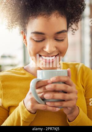 Junge, fröhliche Mischrassen-Frau, die zu Hause eine Tasse Kaffee hält und trinkt. Eine gemütliche hispanische Frau genießt eine Tasse Tee, während sie sich zu Hause entspannt Stockfoto