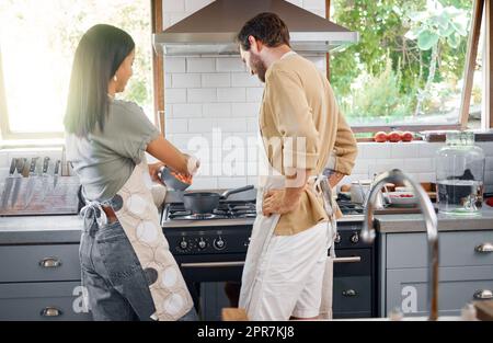 Rückansicht eines jungen gemischtrassigen Paares, das zu Hause eine gesunde Mahlzeit kocht. Fröhliche junge hispanische Frau, die ihrem Mann das Kochen beibringt, indem sie Gemüse in einen Topf auf dem Herd wirft Stockfoto