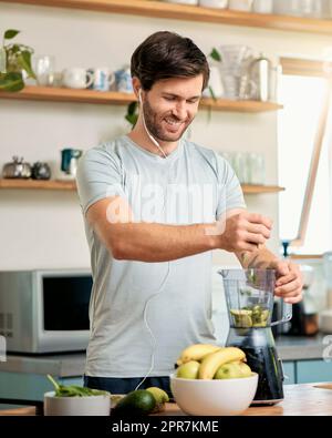 Ein passender junger weißer Mann, der Zutaten in den Mixer gibt, um einen gesunden grünen Entgiftungscreme zu machen, während er zu Hause Kopfhörer in der Küche trägt. Ein Typ, der frischen Fruchtsaft zum Reinigen hat und Energie für das Training bereitstellt. Gesundes Getränk mit Vitaminen und Nährstoffen Stockfoto