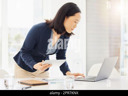 Fokussierte junge asiatische Geschäftsfrau, die ein digitales Tablet und ein Notebook verwendet, während sie Notizen am Schreibtisch im Büro plant. Eine Frau surft nur online, während sie Forschungsdokumente plant und Daten prüft. Rechtsanwalt, der Rechtsberichte erstellt Stockfoto