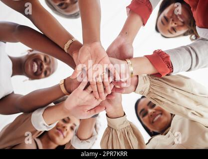 Kleine, vielfältige Gruppe ehrgeiziger, lächelnder Geschäftsfrauen, zusammengedrängt mit Händen in der Mitte. Lächeln, ethnisches Team aus professionellen Kollegen, die sich motiviert, vereint, unterstützt und bereit fühlen Stockfoto