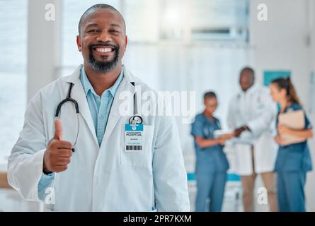 Reifer afroamerikanischer männlicher Arzt zeigt Daumen nach oben, während er mit Kollegen in einem Krankenhaus arbeitet. Fachkundiger Arzt lächelt während einer Handbewegung zur Unterstützung bei der Arbeit in einer Klinik Stockfoto