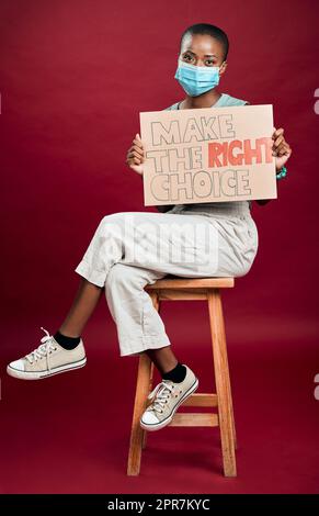 afroamerikanische covid geimpfte Frau zeigt, hält Plakat und trägt chirurgische Gesichtsmaske. Happy schwarz Modell isoliert auf roten Studio Hintergrund wi Stockfoto