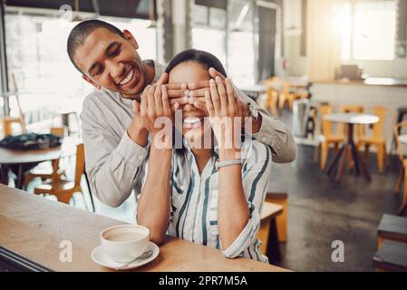 Ein fröhlicher junger Mann, der seine Freundinnen in die Augen hält und sie überrascht, während er in einem Café sitzt. Ein glückliches junges gemischtes Paar, das sich zum ersten Mal zum Kaffee trifft. Aufgeregte Frau versucht zu erraten, wer hinter ihr steckt Stockfoto