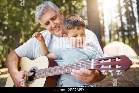 Ein hübscher reifer Mann, der seinem Enkel beibringt, wie man Gitarre spielt, während er im Wald zeltet. Stockfoto