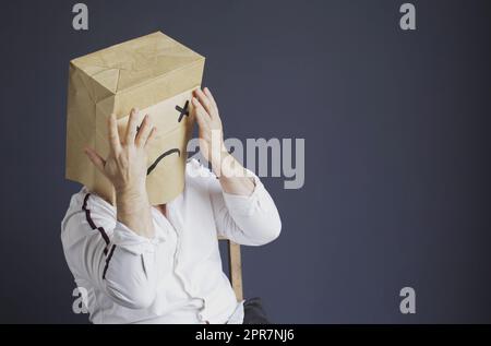 Ein trauriger Mann in einem weißen Hemd mit einer Tasche auf dem Kopf, mit einem gezeichneten traurigen Emoticon, hat Angst und schließt seine Augen mit seinen Händen. Emotionen und Gesten. Stockfoto