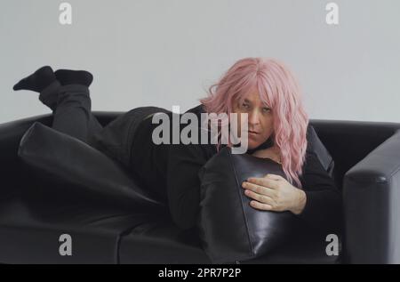 Ein betrunkener Mann mit einer rosa Perücke liegt auf der Couch, wacht auf, hebt seinen Kopf und glättet seine Haare. Stockfoto