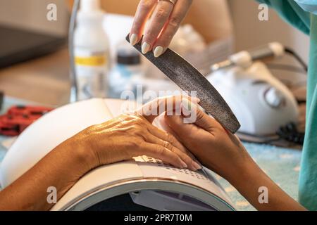 Maniküre der Hände. Nahaufnahme der Hände einer professionellen Maniküre, die die Nägel einer Frau feilen. Verschwommener Hintergrund des Nagelsalons. Nageldesign. Makro. Stockfoto
