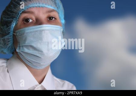 Nahaufnahme des Porträts einer Ärztin in einer medizinischen Mütze und Maske gegen den blauen Himmel. Stockfoto