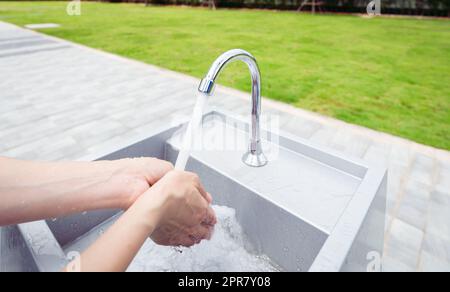 Frau wäscht sich die Hände mit Leitungswasser unter Wasserhahn am weißen Waschbecken. Waschen Sie die Hände mit Leitungswasser am Waschbecken im Freien in der Nähe des Grasfeldes. Persönliche Hygiene zur Vorbeugung von Coronavirus oder Covid 19. Gesunder Lebensstil. Stockfoto