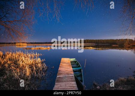 Weißrussland, Osteuropa. Echte Nachthimmel Stars Über Dem Alten Pier Mit Verankertem Hölzernen Fischerboot. Natürlicher Sternenhimmel Und Landschaftslandschaft Mit Lake River Am Frühen Frühlingsabend. Russische Natur Stockfoto