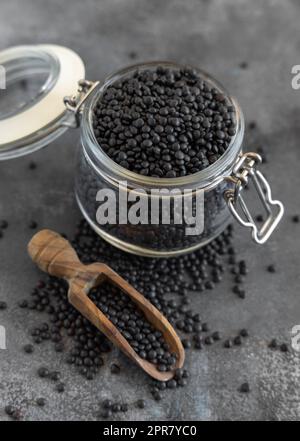 Glasbehälter aus trockenen schwarzen Linsen Bohnen mit einer Schaufel auf grauem Tisch Nahaufnahme, gesunde Protein-Diät Stockfoto