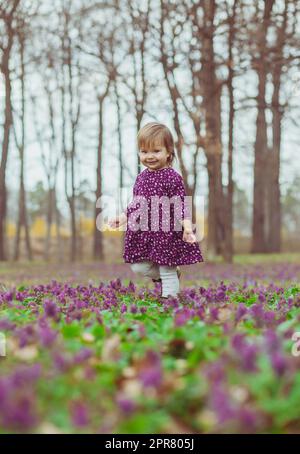 Blondes Baby in einem farbigen Kleid läuft in einer Waldlude mit Blumen Stockfoto