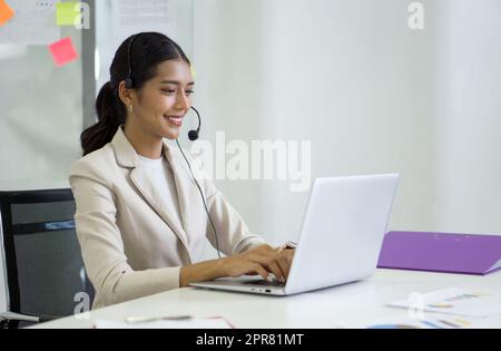 Junge asiatische Geschäftsfrau in braunem Anzug und Headset mit Mikrofon, die auf der Tastatur des Laptops tippt, während sie sich online über eine Internetverbindung trifft. Arbeitsbegriff soziale Distanzierung. Stockfoto