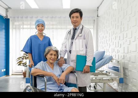 Ältere Patientin und Ärztin. Die junge Krankenschwester schiebt einen Rollstuhl des älteren Patienten, während ein asiatischer Arzt mit Stethoskop und Kleid daneben stand. Stockfoto