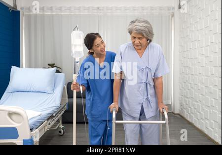 Junge asiatische Krankenschwester im Pflegeheim pflegen behinderte Seniorin. Der behandelnde Arzt bietet eine Physiotherapie für ältere Patienten an, um auf Gehhilfe oder Gehstock zu trainieren. Stockfoto