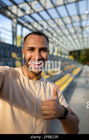 Nahaufnahme. Vertikales Foto. Junger afroamerikanischer Sportler steht im Stadion mit Kopfhörern, macht ein Selfie am Telefon, spricht bei einem Videoanruf. Zeigt der Kamera eine Super-Geste an, Stockfoto