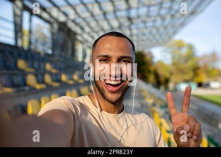 Nahaufnahme. Junger afroamerikanischer Sportler, der im Stadion in Kopfhörern steht und ein Selfie am Telefon macht. Er zeigt es der Kamera mit seinen Fingern als Zeichen des Sieges, lächelt. Stockfoto