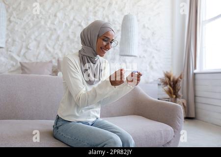 Foto von der Seite. Lächelnde junge muslimische Frau im Hijab, die zu Hause auf dem Sofa sitzt und Einweg- und Schnellschwangerschaftstest hält. Zufrieden mit dem positiven Ergebnis. Stockfoto