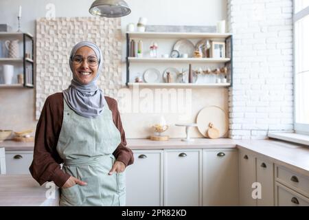 Porträt einer jungen arabischen Hausfrau im Hidschab, die in der Küche neben dem Tisch in der Schürze steht, Hände in den Taschen hält, lächelt und in die Kamera schaut. Stockfoto