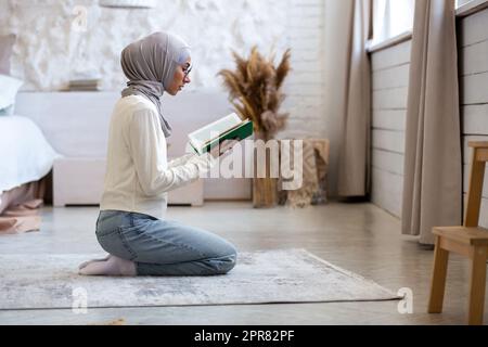 Eine junge Frau orientalischer Herkunft und Religion sitzt in einem Hidschab zu Hause auf ihren Knien und mit gefalteten Händen, schließt ihre Augen und betet. Er hält ein heiliges Buch in seinen Händen, liest ein Gebet. Stockfoto