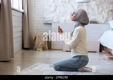Eine junge muslimische Frau in einem Hidschab sitzt zu Hause auf ihren Knien und faltete ihre Hände, hob ihren Kopf hoch, blickt aus dem Fenster und betet. Stockfoto