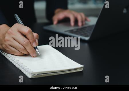 Ein Geschäftsmann hält eine Buchnotiz mit Stift auf dem Schreibtisch und einem Computer mit Laptop. Stockfoto