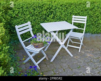 Zwei Innenhofstühle und Tisch in einem abgeschiedenen und privaten Garten zu Hause. Holzmöbel und Sitzbereich für Kaffee und Lesen in einem Hinterhof. Lila Wiese, Cranesbill-Blumen und eine Hecke Stockfoto