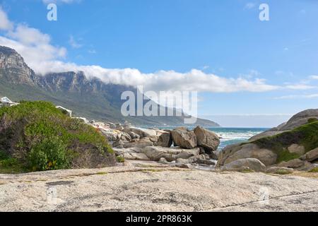 Wunderschöne Landschaft von Camps Bay in Kapstadt, Südafrika. Malerische Berge und Felsen in der Nähe des Ozeans oder des Meeres mit blauem Hintergrund. Friedliche Natur am Strand mit Meereslandschaft an einem Sommertag Stockfoto