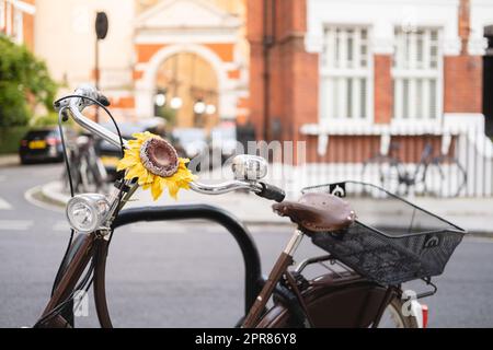 Horizontale Nahaufnahme eines Fahrrads in braunem Vintage-Look mit einer gelben Sonnenblume am Lenker. Das Fahrrad hat eine Glocke, Lichter und einen Korb, ist parke Stockfoto