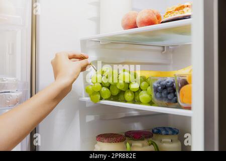 Eine Frau, die Trauben aus einem offenen Kühlschrank oder Kühlschrank nimmt oder aufnimmt Stockfoto