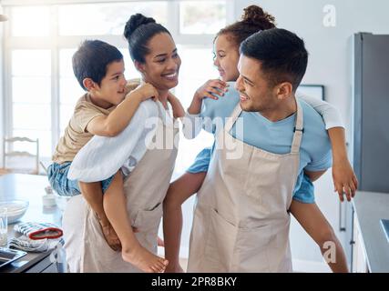 Energiegeladene Eltern fahren ihre Kinder mit dem Huckepack, während sie Schürzen tragen und in der Küche backen. Hispanische eltern spielen zu Hause mit Kindern Stockfoto