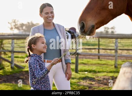 Hey, horsey, ich hab dir einen Snack. Ein entzückendes kleines Mädchen füttert ein Pferd auf ihrer Farm, während ihre Mutter auf sie schaut. Stockfoto