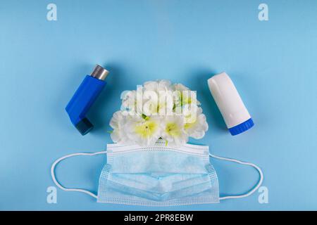 Zwei blaue Inhalatoren, medizinische Maske und weiße Blumen auf blauem Hintergrund mit negativem Platz für die Platzierung von Kopien. Atemsensibilität Asthma-Konzept Stockfoto